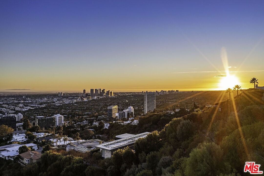 This stunning aerial perspective captures a panoramic sunset view over the Los Angeles skyline, highlighting the expansive urban landscape and distant horizon. In the foreground, a modern architectural residence with a flat roof is nestled into the lush, hillside terrain, emphasizing its prime location and elevated vantage point. The golden hour light casts a warm, dramatic glow across the city, creating a sophisticated and highly desirable atmosphere.