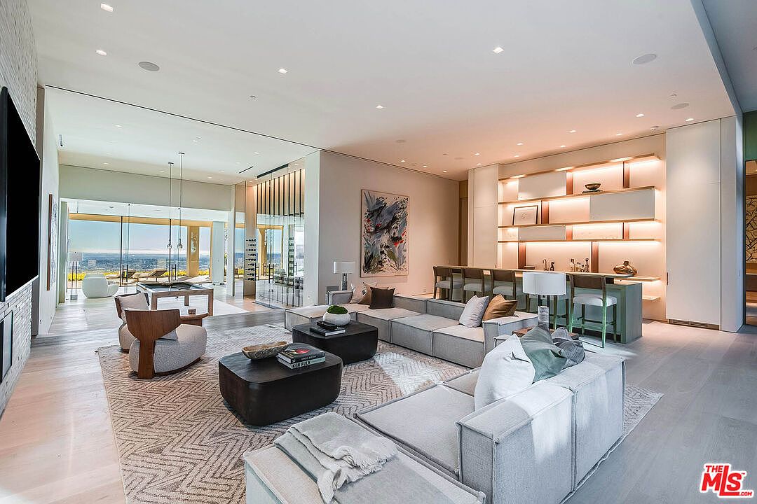 This expansive, open-concept living room exudes modern luxury with its neutral color palette, plush sectional seating, and sophisticated lighting design. The space seamlessly transitions into a bar area and a sun-drenched lounge with panoramic views, highlighting a high-end, contemporary aesthetic perfect for entertaining. The clean lines, textured area rug, and curated artwork create a refined and inviting atmosphere.