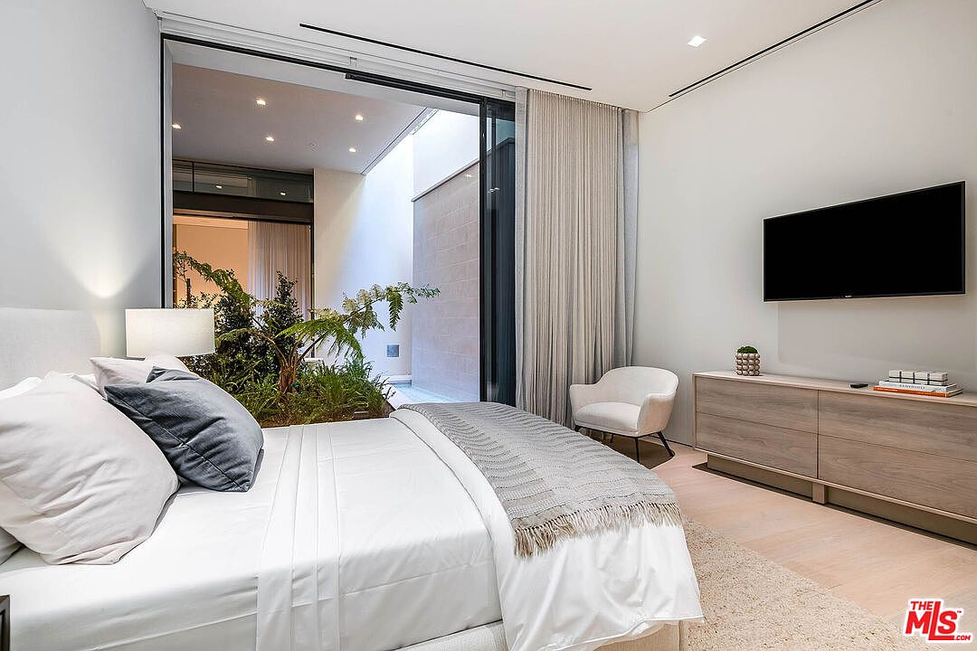 This modern primary bedroom features a minimalist aesthetic with a plush bed, a sleek wooden dresser, and a wall-mounted television. A large floor-to-ceiling glass door opens to a private, lush indoor-outdoor courtyard garden, creating a serene and tranquil atmosphere. The neutral color palette and clean lines emphasize a sophisticated, high-end contemporary design.