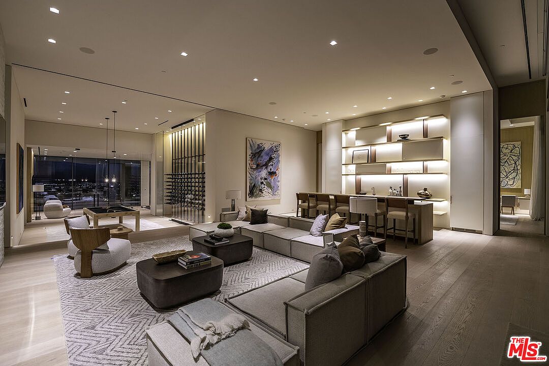 This expansive, modern living room features a sophisticated open-concept design with a large sectional sofa, plush armchairs, and dual coffee tables arranged on a textured area rug. The space is anchored by a sleek, illuminated bar area with integrated shelving and a glass-enclosed wine display, creating a luxurious atmosphere perfect for entertaining. Large floor-to-ceiling windows provide a seamless transition to an outdoor terrace, while recessed lighting and contemporary artwork enhance the refined, high-end aesthetic.