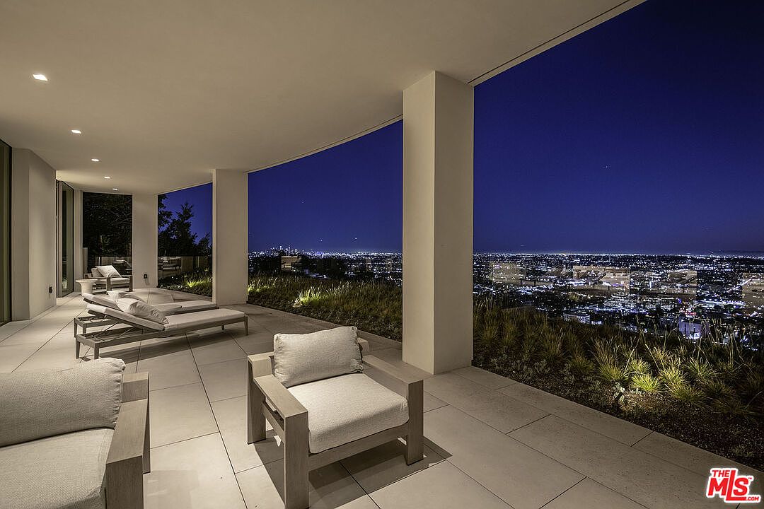 This expansive, modern covered balcony offers a breathtaking panoramic view of the city lights at night. The space is furnished with sleek, contemporary lounge chairs and chaise lounges, set against a backdrop of minimalist architecture and integrated landscape lighting. The open-air design creates a sophisticated and serene atmosphere, perfect for evening relaxation or entertaining.