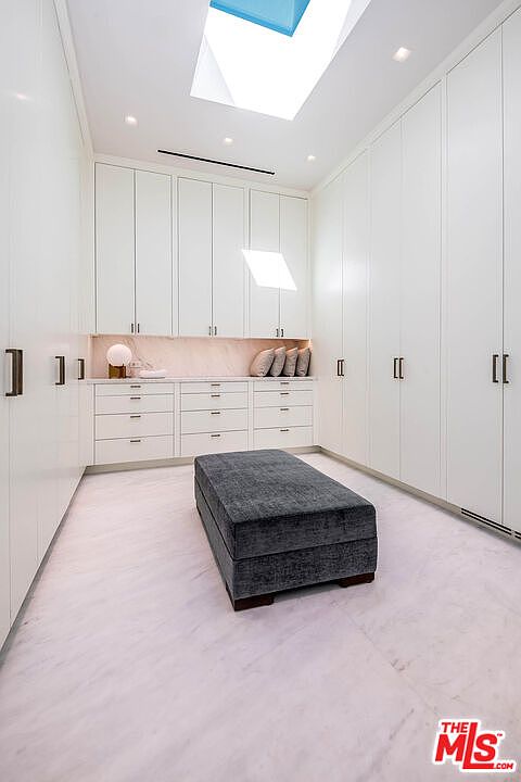 This luxurious walk-in closet features floor-to-ceiling white cabinetry, a central dark grey upholstered ottoman, and elegant marble flooring. A large skylight overhead floods the space with natural light, highlighting the clean, minimalist aesthetic and sophisticated storage solutions. The perspective is centered, showcasing the symmetrical layout and high-end finishes typical of a modern primary suite.