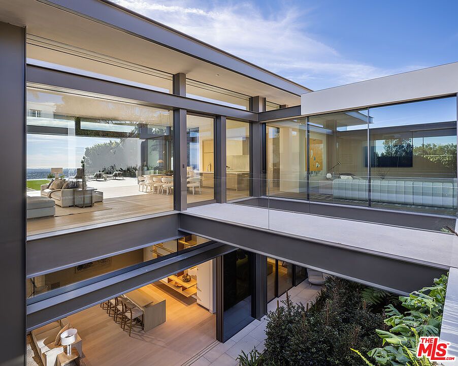 This striking architectural shot captures a multi-level modern residence featuring an open-air bridge connecting two wings of the home. The design emphasizes indoor-outdoor living with expansive floor-to-ceiling glass walls, sleek steel structural elements, and a lush, sunken garden courtyard visible below. The perspective offers a sophisticated, cinematic view of the home's contemporary aesthetic and seamless integration with the surrounding landscape.