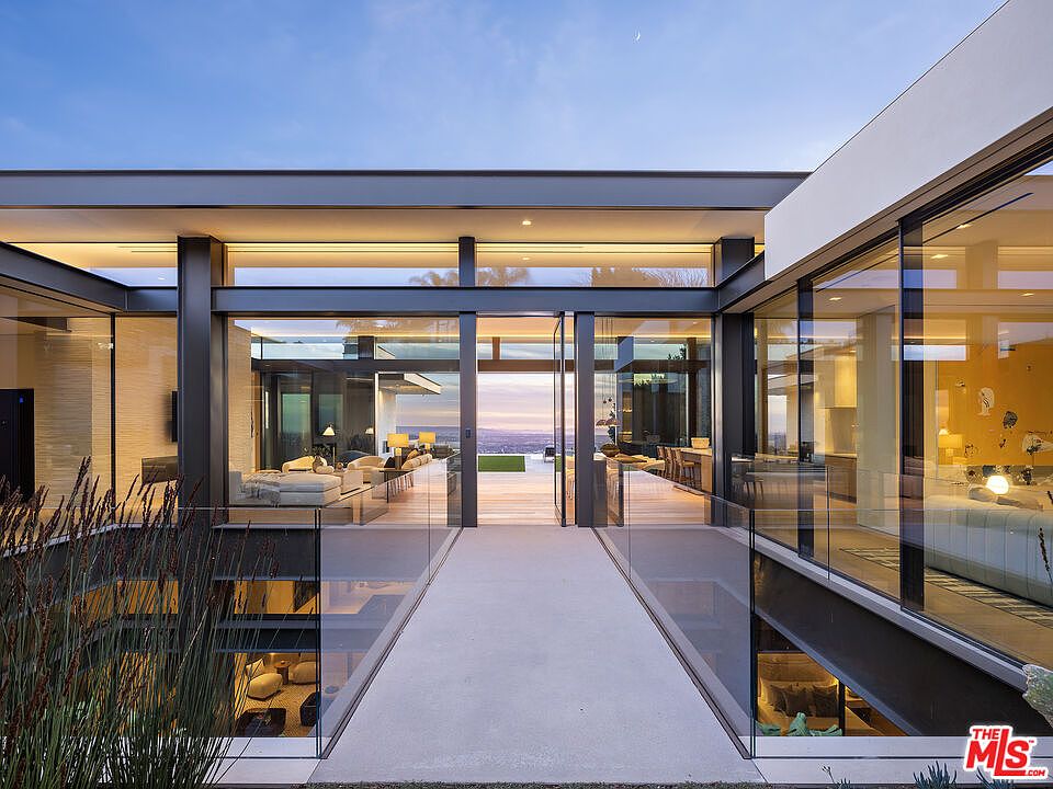 This striking modern entryway features a minimalist concrete bridge leading directly into an expansive, open-concept living space framed by floor-to-ceiling glass walls. The architectural design emphasizes seamless indoor-outdoor integration, showcasing a sophisticated steel structure and a panoramic view of the horizon at dusk. The perspective is centered and symmetrical, highlighting the clean lines and luxurious, contemporary aesthetic of the residence.