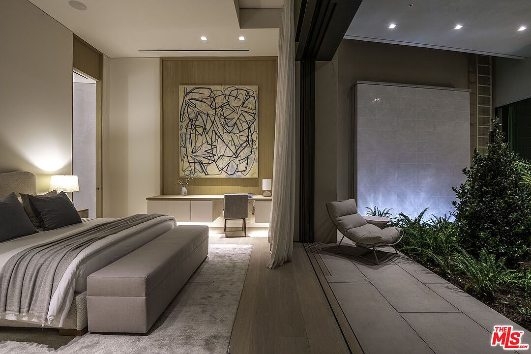 This luxurious primary bedroom features a seamless transition between a sophisticated sleeping area and an indoor-outdoor garden space. The room is highlighted by a minimalist aesthetic, a large abstract art piece above a built-in desk, and a serene water feature wall adjacent to lush greenery. The open-concept design, combined with soft ambient lighting and neutral tones, creates a tranquil and high-end atmosphere.