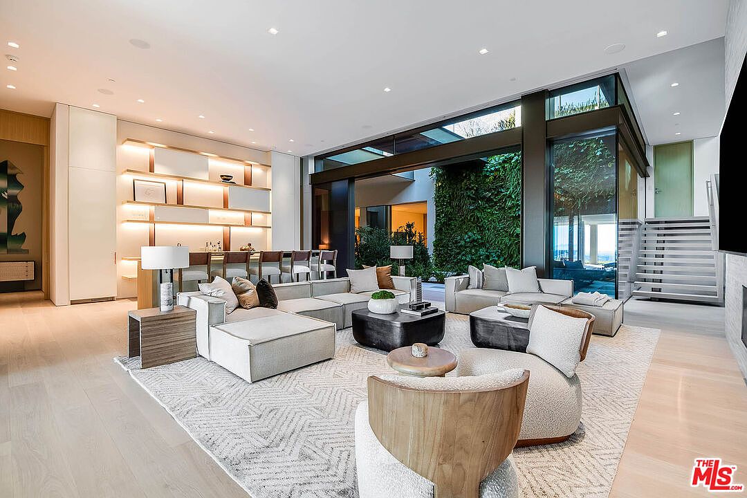 This expansive, modern living room features a sophisticated open-concept design with high ceilings and floor-to-ceiling glass walls that seamlessly connect to an indoor-outdoor courtyard with a lush green wall. The space is furnished with plush, neutral-toned sectional sofas, a sculptural armchair, and a sleek coffee table, all anchored by a large, textured area rug. A built-in bar area with illuminated shelving adds a touch of luxury, creating an ideal environment for both relaxation and high-end entertaining.