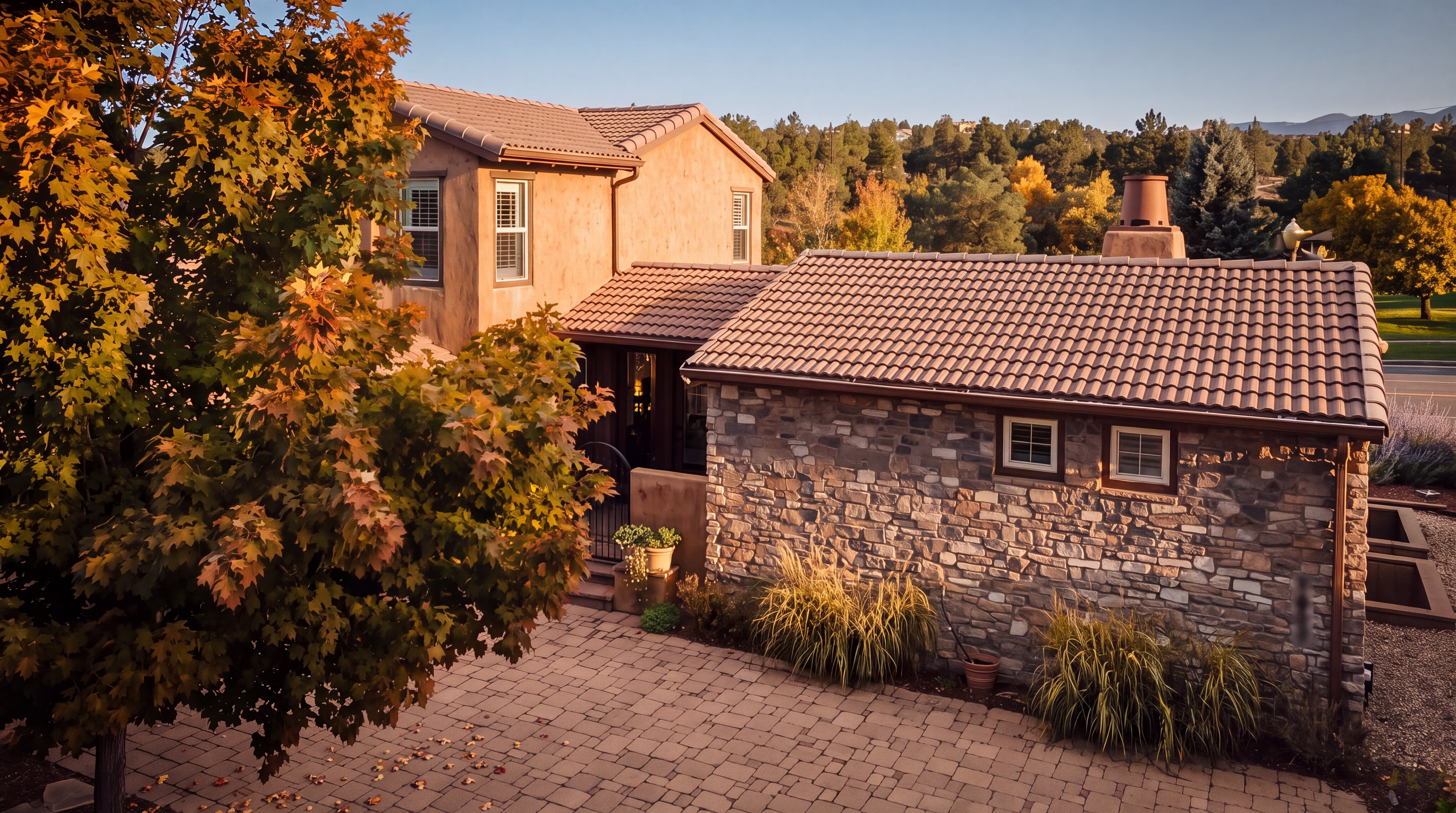 This side view captures a multi-level home featuring a combination of textured stucco walls and a stone-clad exterior section. A paved stone patio area sits in the foreground, framed by a large, leafy tree and ornamental grasses. The perspective highlights the architectural transition between the two building materials and the tiled roofline against a clear blue sky.