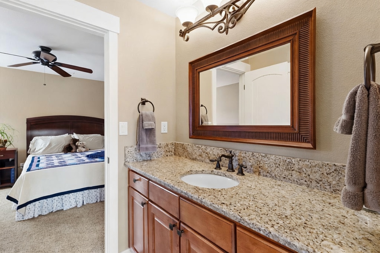 This bathroom features a vanity with a stone countertop, an oval undermount sink, and a framed mirror mounted above. To the left, a doorway provides a clear view into an adjacent bedroom, which is furnished with a dark wood bed frame and a ceiling fan. The space is finished with neutral wall tones and warm-toned cabinetry, creating a cohesive and functional transition between the two rooms.