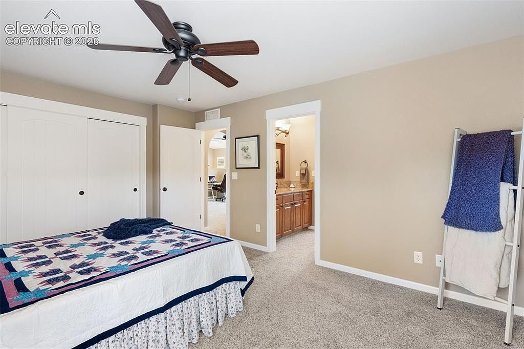 This bedroom features neutral-toned walls, plush carpeting, and a ceiling fan centered above a bed dressed in a patterned quilt. A white closet with sliding doors occupies one wall, while an open doorway leads into an adjoining bathroom with wood-toned cabinetry. A metal ladder-style rack stands against the wall, holding folded blankets, creating a tidy and functional living space.