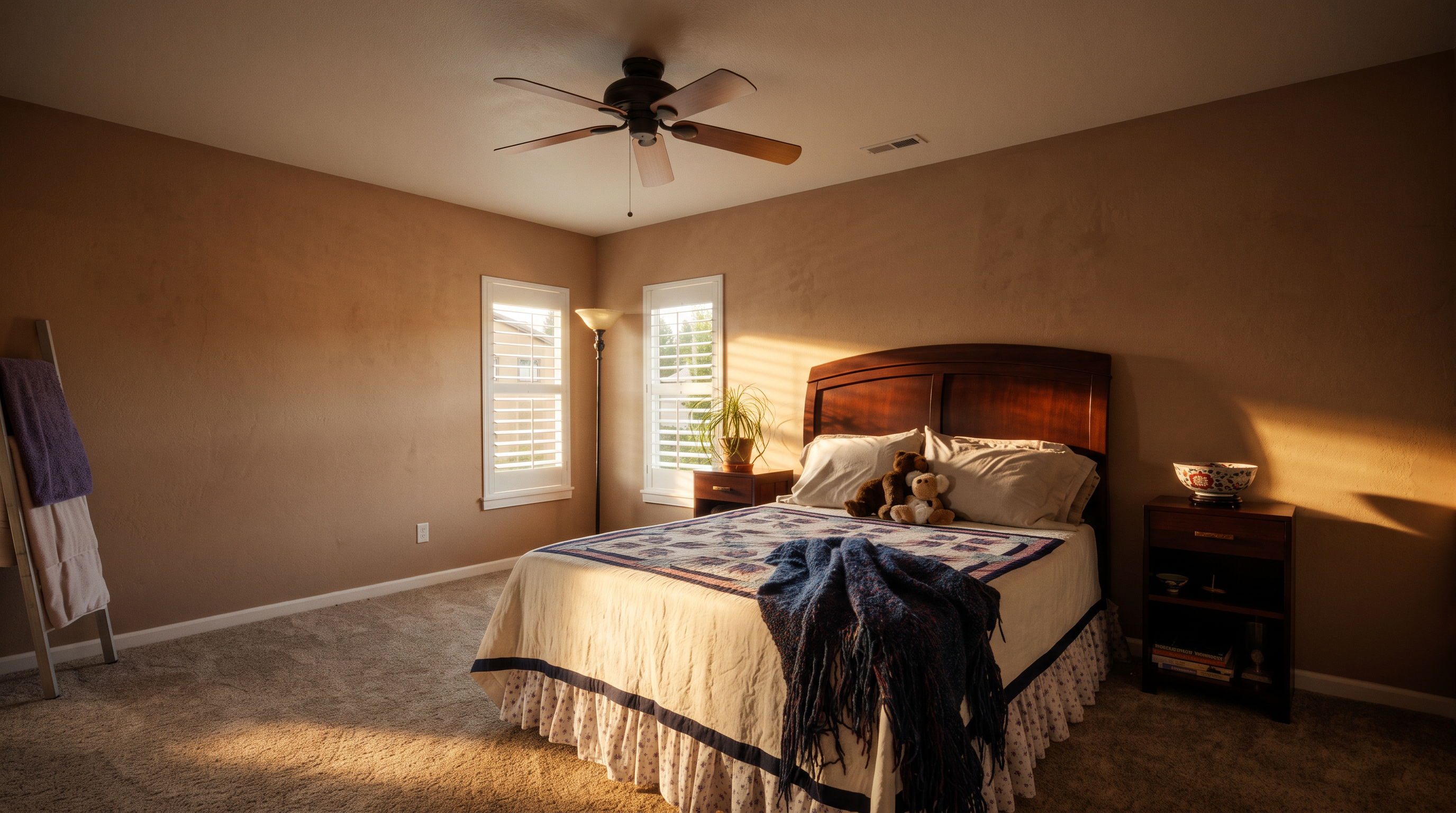 This bedroom features neutral-toned walls and wall-to-wall carpeting, creating a calm and inviting atmosphere. The room is furnished with a dark wood bed frame topped with a patterned quilt and a matching nightstand, while two windows with white shutters allow for natural light. A ceiling fan with a light fixture is centered above, and a floor lamp stands in the corner, completing the functional layout.