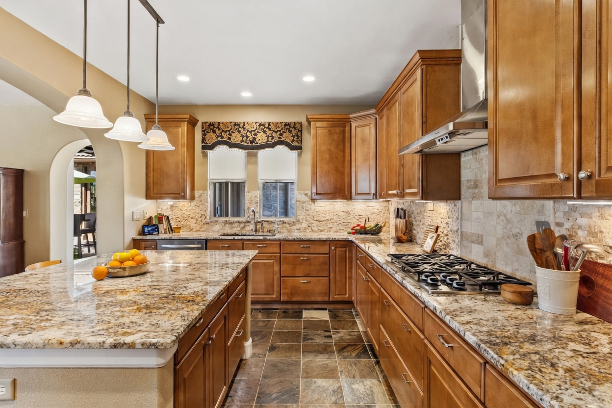 This spacious kitchen features warm-toned wood cabinetry, a large central island with a stone countertop, and a gas cooktop with a stainless steel range hood. The room is accented by a decorative valance over the sink window and pendant lighting, creating a welcoming and functional culinary space. The perspective is a wide-angle shot from the kitchen island looking toward the main cooking area, showcasing the stone backsplash and tiled flooring.