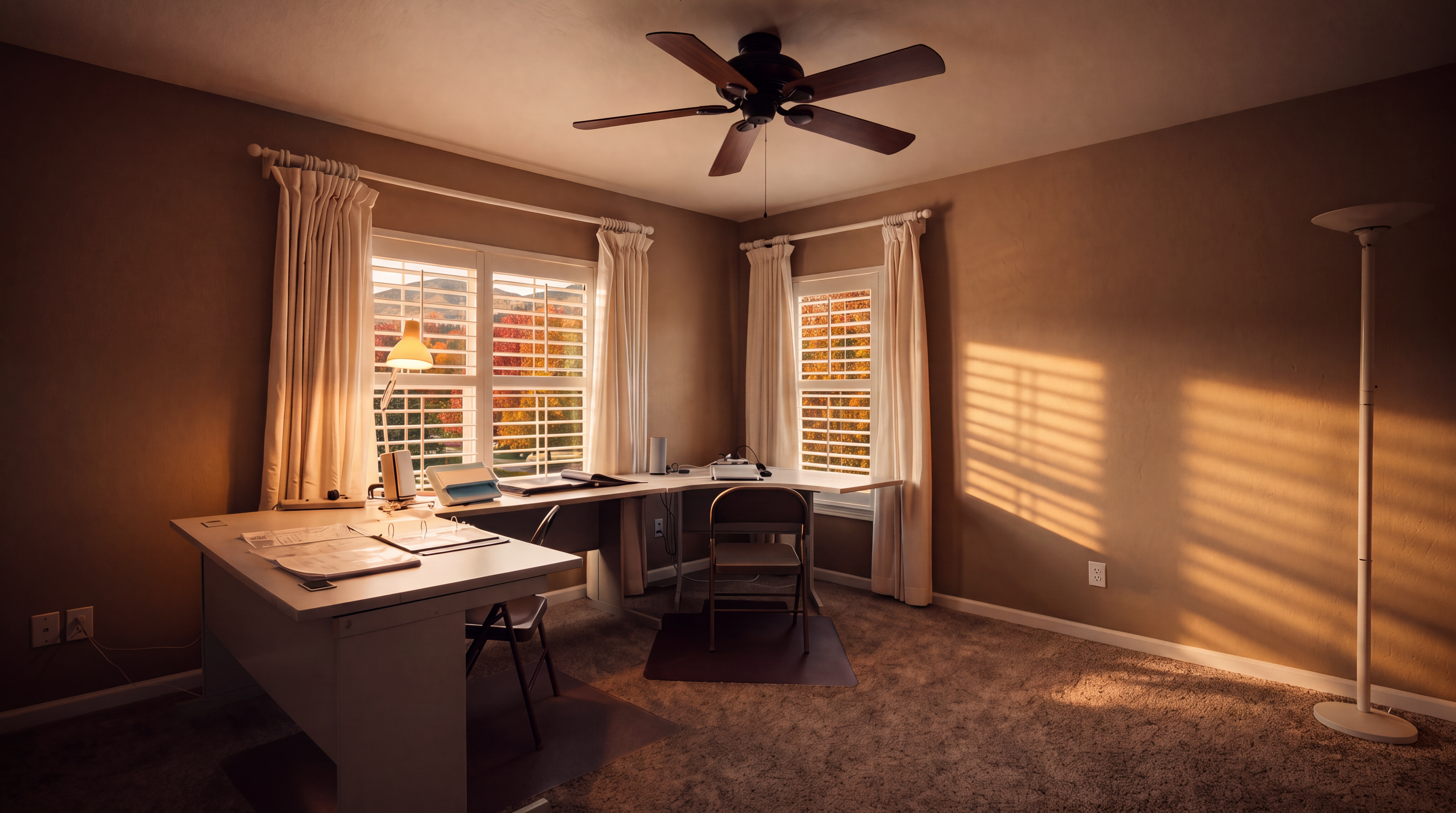 This home office features neutral-toned walls and wall-to-wall carpeting, creating a professional and quiet workspace. A large, L-shaped desk is positioned in the corner, complemented by two chairs and illuminated by a ceiling fan with integrated lighting. Large windows with white curtains allow natural light to brighten the room, while a tall floor lamp provides additional task lighting.