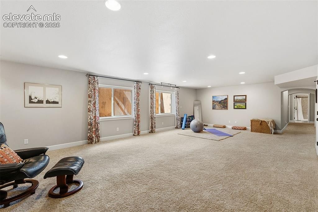 This spacious, carpeted basement area offers a versatile layout suitable for a home gym or recreational space. The room features neutral walls, two sets of windows with patterned curtains, and recessed lighting throughout. A yoga mat, exercise ball, and a woven storage basket are positioned in the center, while a comfortable leather armchair and ottoman occupy the foreground.