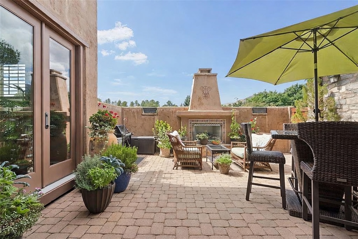 This inviting outdoor patio features a paved brick floor and a prominent stucco-finished fireplace as its central focal point. The space is furnished with wicker-style seating and a high-top table under a lime green umbrella, creating a comfortable area for relaxation or dining. The scene is framed by potted plants and a low wall, offering a private and serene atmosphere under a clear blue sky.