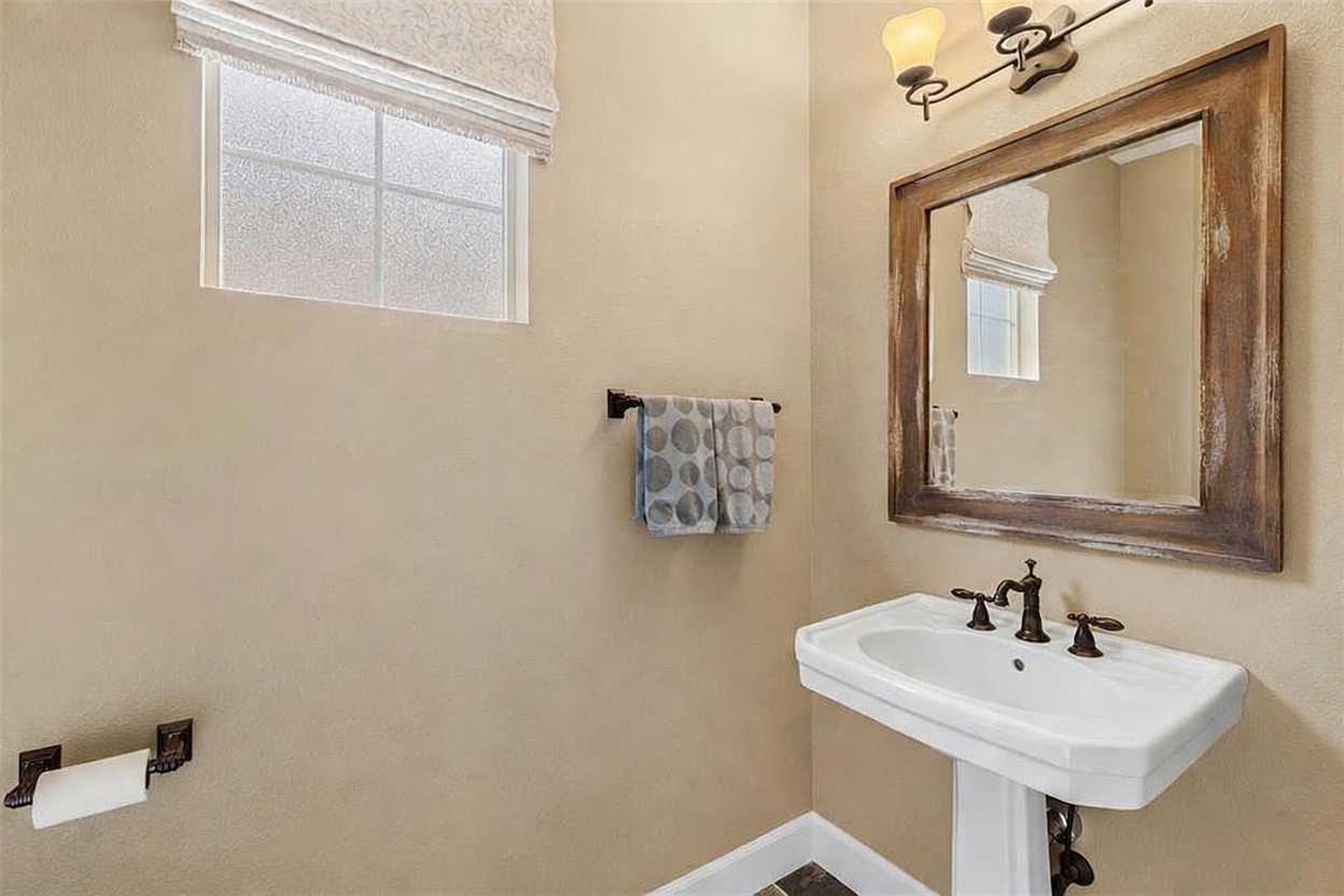 This compact powder room features a classic pedestal sink with a dark-finished faucet and a matching framed mirror above. A patterned hand towel hangs on a wall-mounted rack, while a toilet paper holder is positioned conveniently to the left. The space is illuminated by a multi-light vanity fixture and natural light filtering through a window with a fabric shade.
