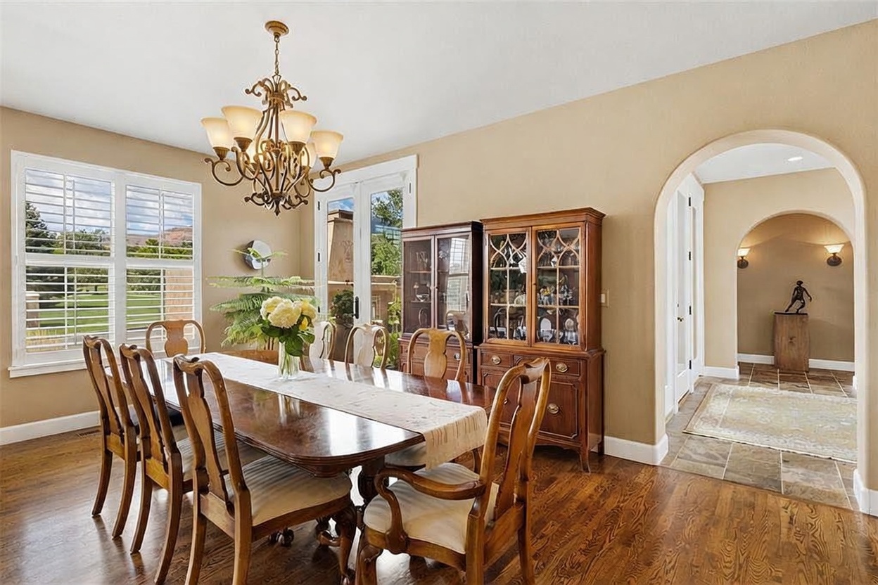 This formal dining room features a large wooden table surrounded by matching chairs, set beneath an ornate, multi-armed chandelier. A tall wooden hutch with glass-front cabinets provides elegant storage, while a wide arched doorway leads into a tiled hallway with a decorative statue. The space is finished with warm-toned walls and rich wood flooring, creating a sophisticated and inviting atmosphere.
