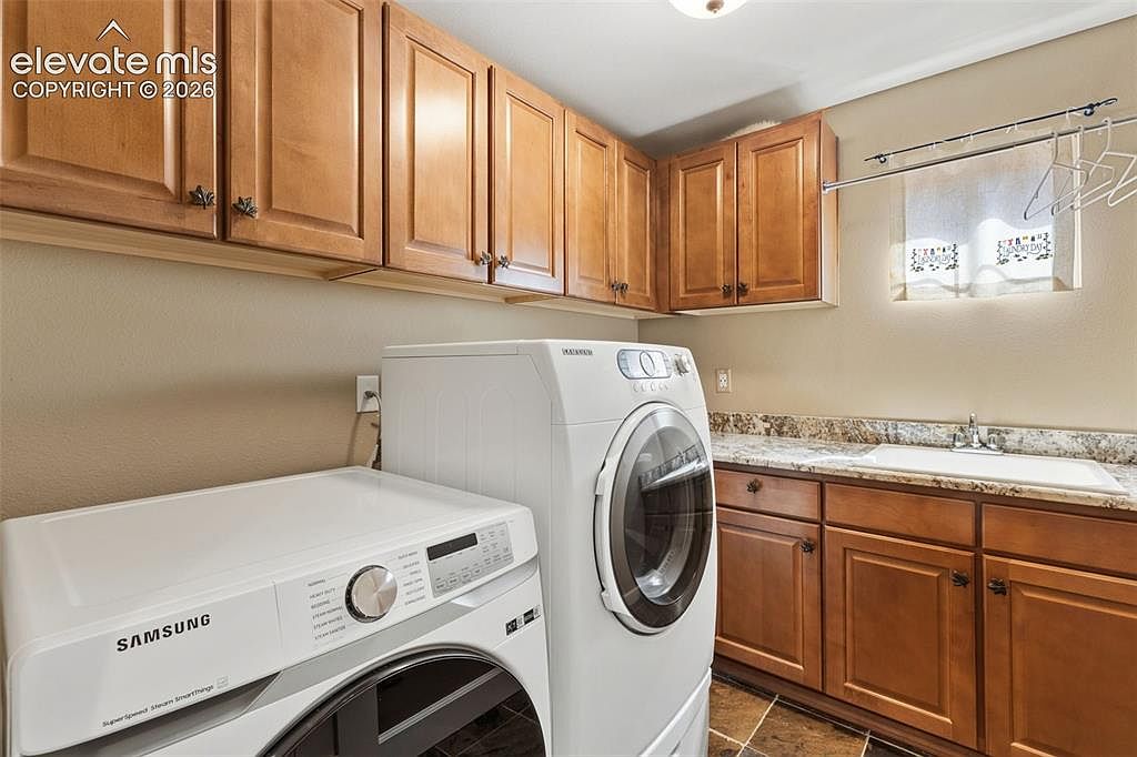 This functional laundry room features a pair of white front-loading Samsung appliances positioned beneath a row of light wood upper cabinets. To the right, a stone-topped counter includes an integrated sink, complemented by matching lower cabinetry and a wall-mounted drying rack. The space is finished with neutral wall tones and tiled flooring, creating a clean and organized utility area.