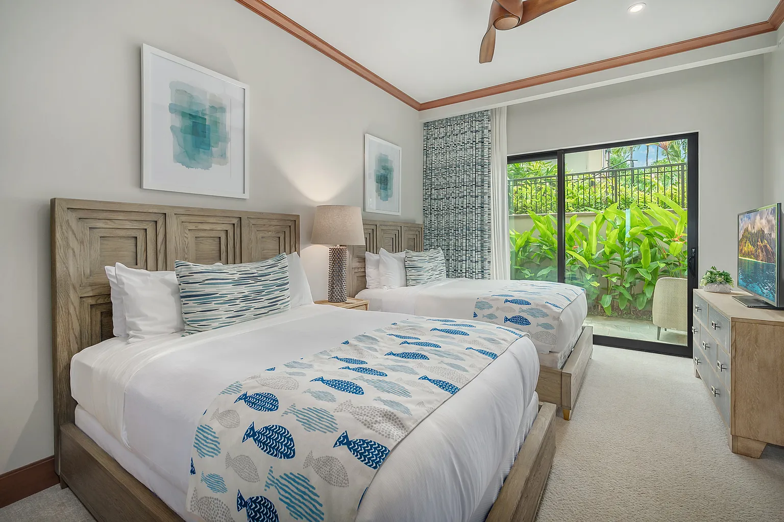 This is a well-lit guest bedroom with two twin beds, neutral walls, and stylish accents. The beds feature wooden headboards, white linens, and blue patterned throws and pillows. A patio door offers access to outdoor greenery, enhancing the room's appeal.