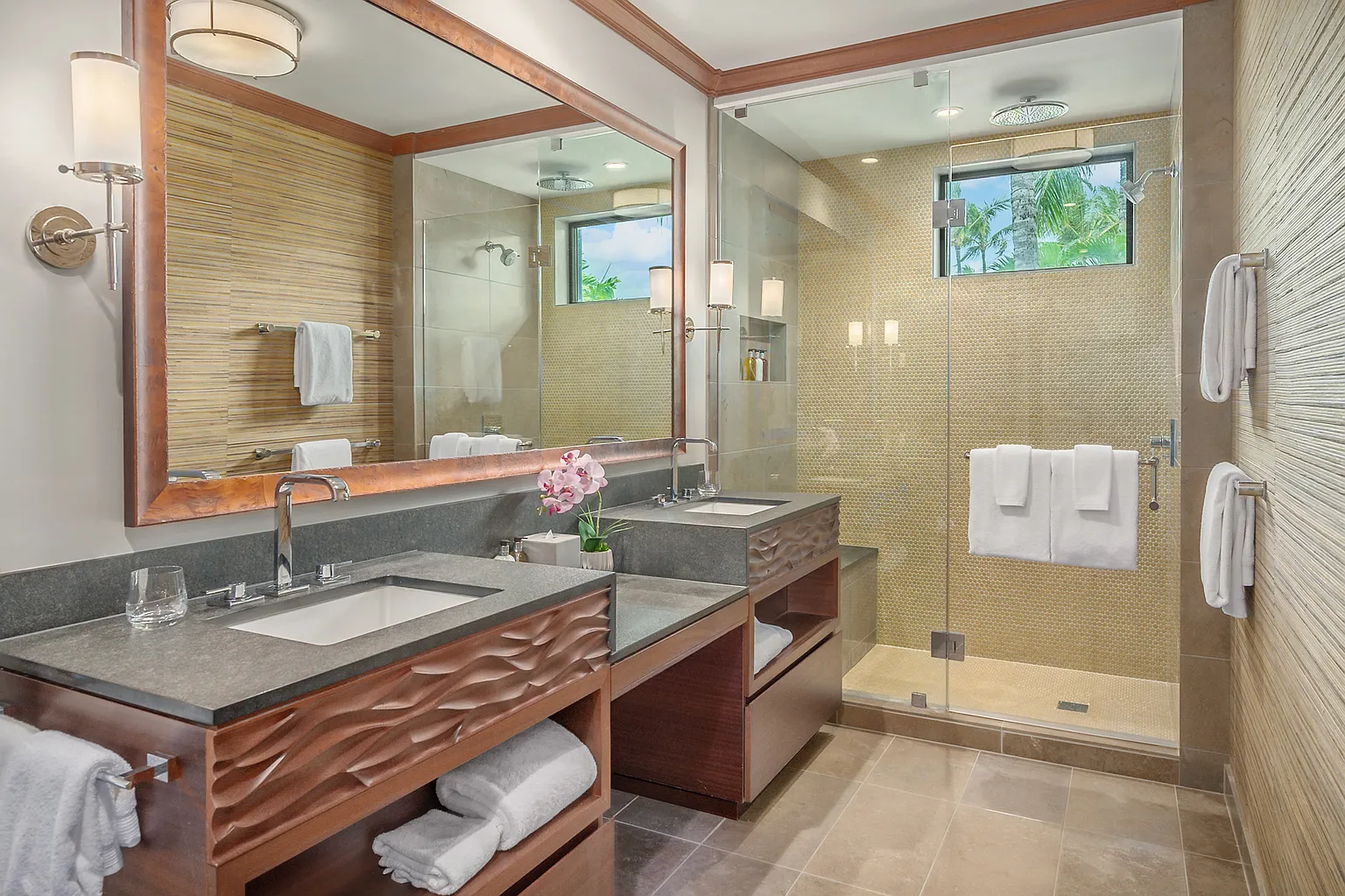 This is a well-designed primary bathroom featuring dual vanities with modern sinks and dark countertops atop custom wood cabinetry with interesting wave details. A large mirror enhances the space, reflecting natural light. The shower is enclosed with glass and the tiling and color palette create a relaxing and luxurious atmosphere.