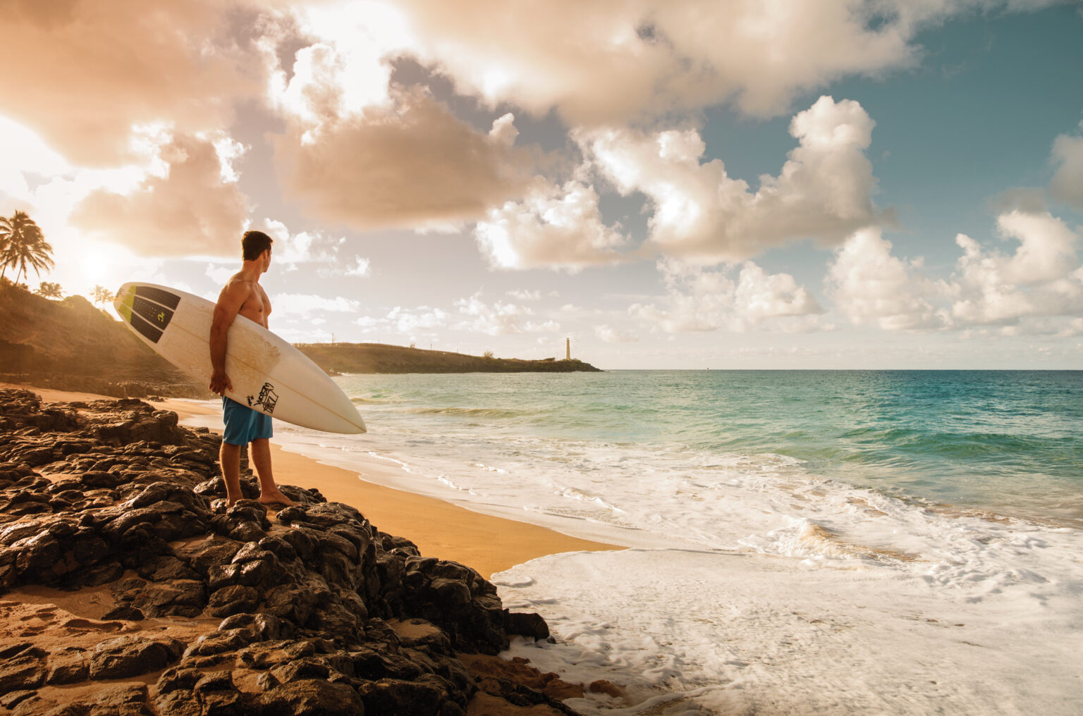 This scenic outdoor shot depicts a male figure holding a surfboard on a rocky outcropping overlooking an ocean beach. The golden sand beach, turquoise waters, and partly cloudy sky create a tranquil and inviting atmosphere. In the background, a distant lighthouse and lush vegetation add depth and interest to the landscape showcasing the property's proximity to natural amenities and recreational opportunities.