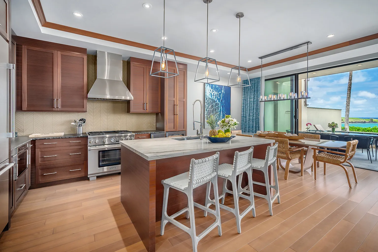 This is a bright and modern kitchen featuring custom wood cabinetry and a contrasting island with seating. Stainless steel appliances and pendant lighting add to the upscale feel, while large sliding glass doors open to an outdoor dining area with ocean views. The hardwood floors throughout create a cohesive and inviting space.