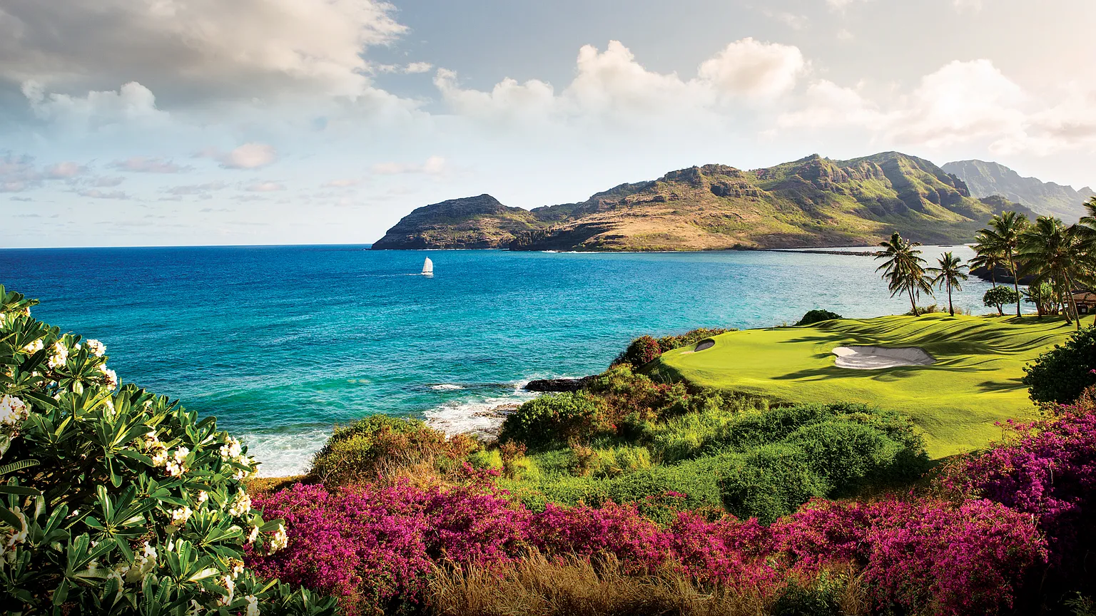 This image showcases a vibrant coastal golf course with stunning ocean views and lush greenery. The foreground features colorful flowering plants, while a well-manicured green with a sand trap sits along the water's edge, framed by palm trees. The turquoise ocean and mountainous backdrop create a picturesque and luxurious setting, perfect for a high-end real estate listing highlighting outdoor amenities and natural beauty.