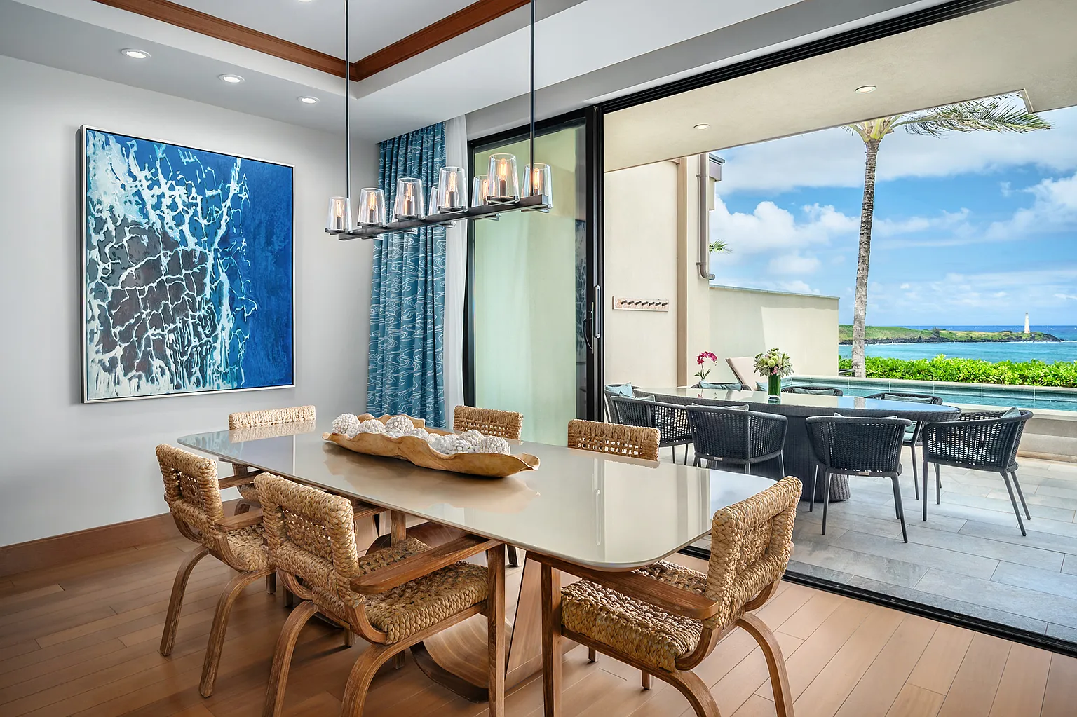 This is a well-lit dining room with modern, luxurious features. The room includes a long dining table with woven chairs, a contemporary chandelier, and a striking blue abstract painting. Sliding glass doors open to an outdoor patio with additional seating and a view of the ocean, blending indoor and outdoor living spaces.