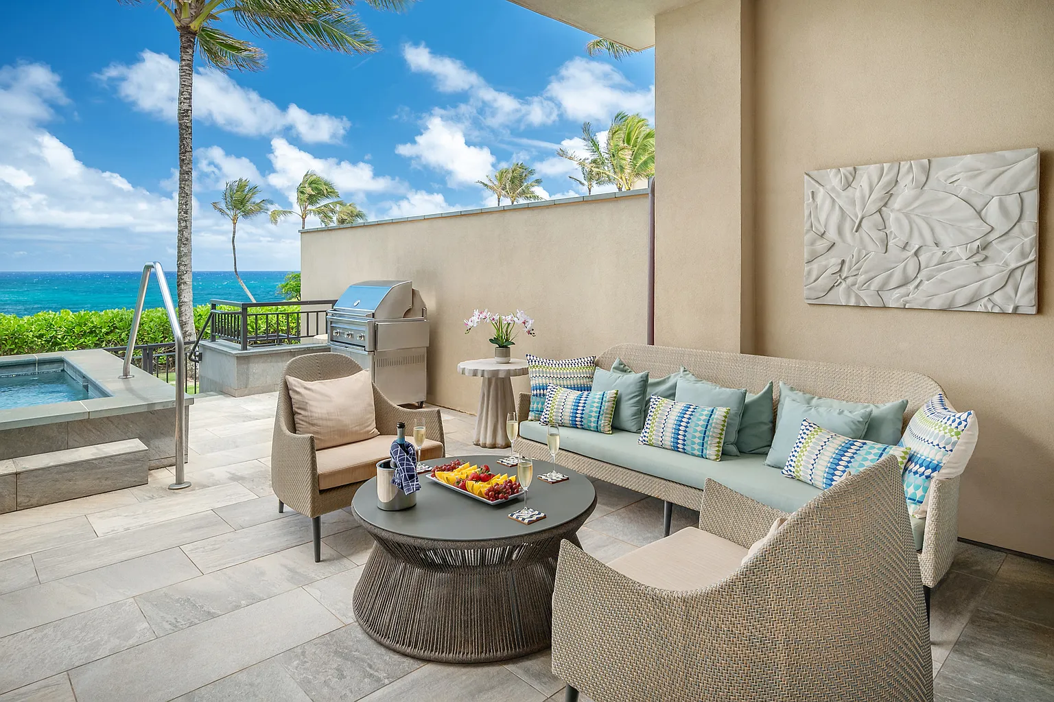 This image showcases a luxurious outdoor living space on a patio or balcony. It features comfortable wicker furniture including a sofa and chairs, stylish throw pillows, and a round coffee table with snacks and beverages. In the background, there is a well-maintained spa, barbecue, and a captivating ocean view, which enhance the overall appeal of this outdoor retreat.
