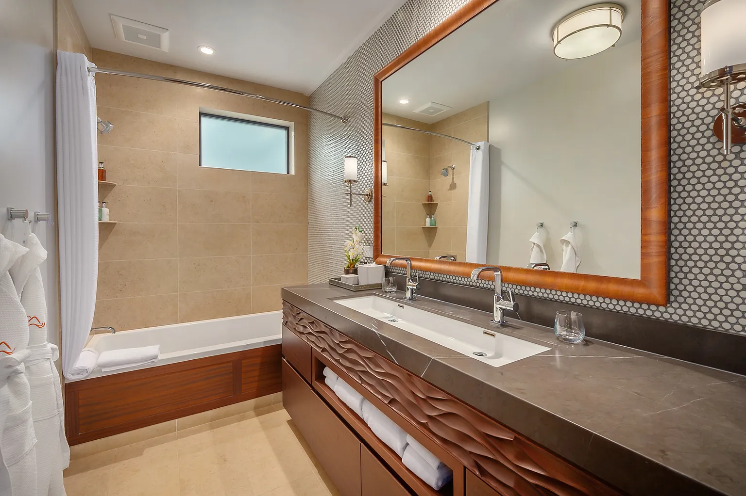 This is a well-lit primary bathroom featuring a double vanity with a dark countertop and decorative wooden cabinetry. A large mirror reflects the space, enhancing its perceived size. The bathroom includes a bathtub with a shower curtain and coordinated tile work, creating a cohesive and upscale aesthetic.