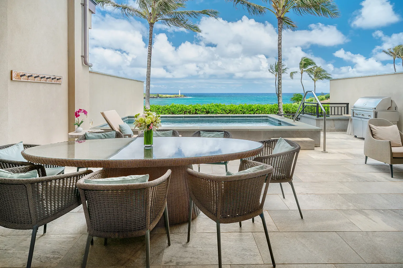 This image showcases an inviting outdoor patio area perfect for relaxation and entertaining. It features a large round table with seating, a swimming pool with ocean views framed by palm trees, and a built-in grill area, creating a luxurious, resort-like atmosphere. The patio's design elegantly marries indoor comfort with outdoor beauty.