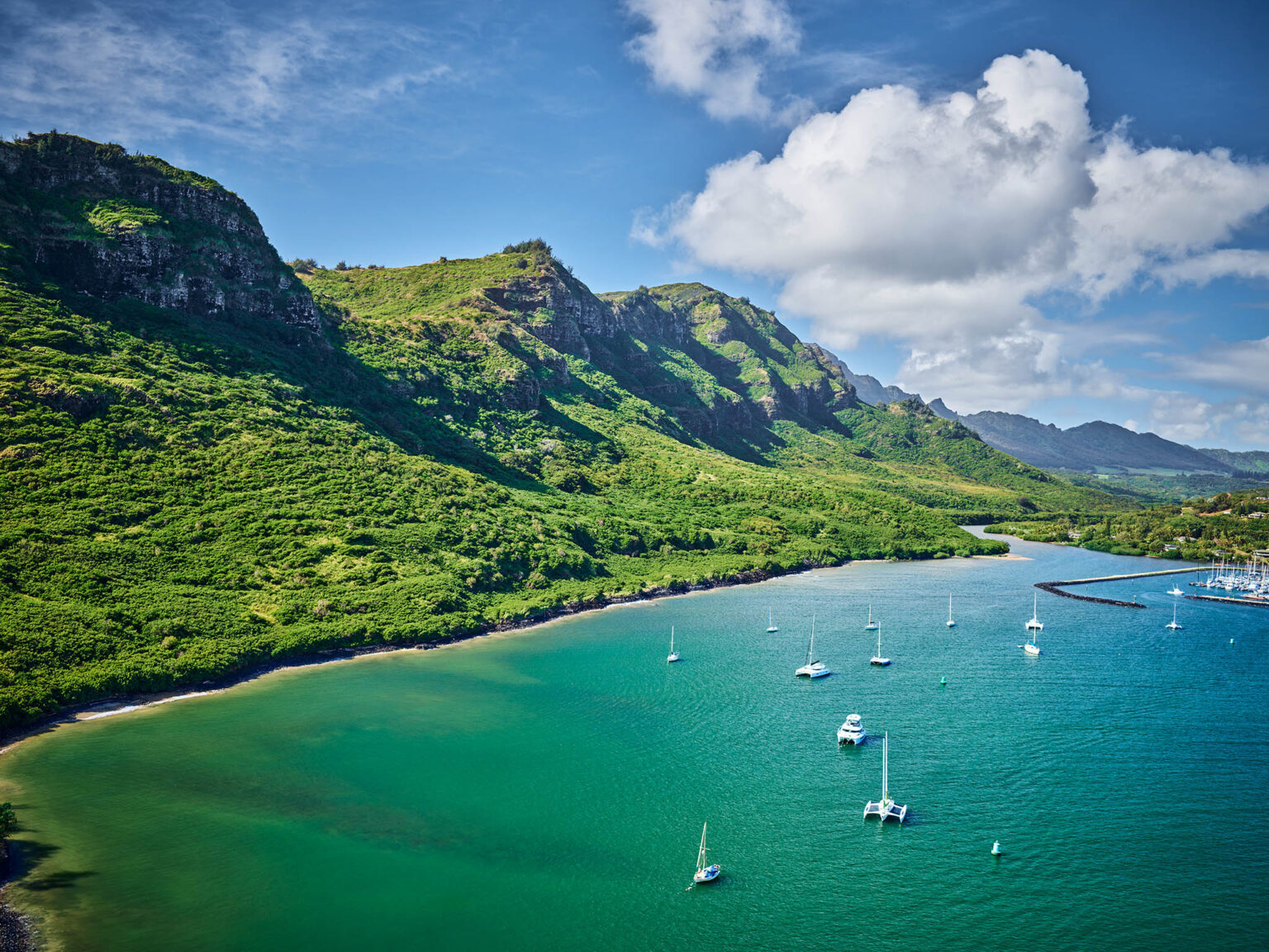 This aerial shot showcases a stunning coastal landscape, potentially highlighting waterfront properties. A lush, green mountain range meets the turquoise ocean waters, where several sailboats and a marina are visible. The scene evokes a sense of tranquility and natural beauty, making it an attractive location for real estate.