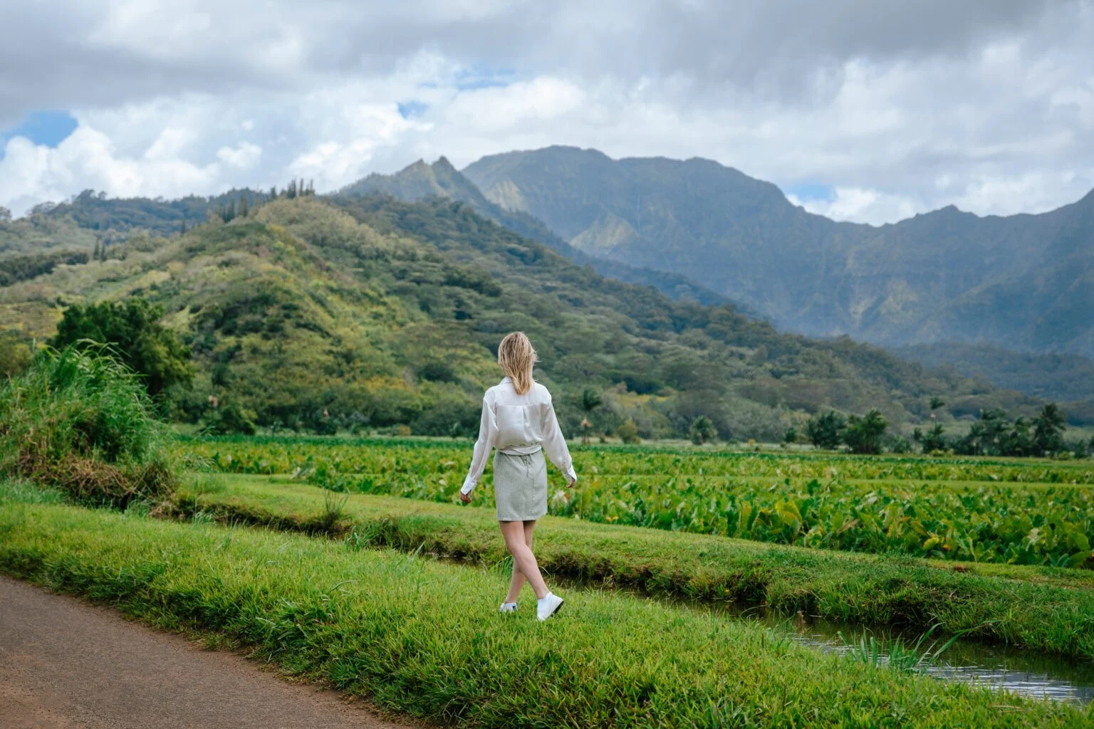 This image showcases an expansive yard with lush greenery and a woman walking through it, suggesting ample outdoor space on the property. The backdrop is a picturesque mountain range, enhancing the property's appeal with a scenic view. A small creek borders the walking path which adds a natural water feature to the landscape.