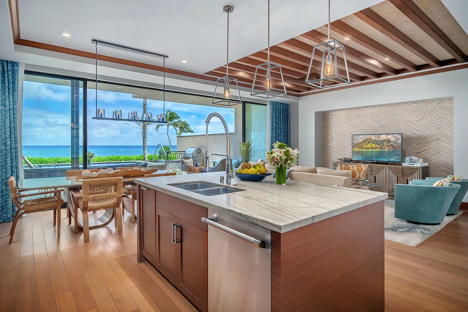 This showcases a modern kitchen with a large center island featuring a white marble countertop and dark wood cabinetry. The kitchen seamlessly flows into the dining and living areas, offering stunning ocean views through expansive windows. Pendant lighting and strategic ceiling design add architectural interest to the space.