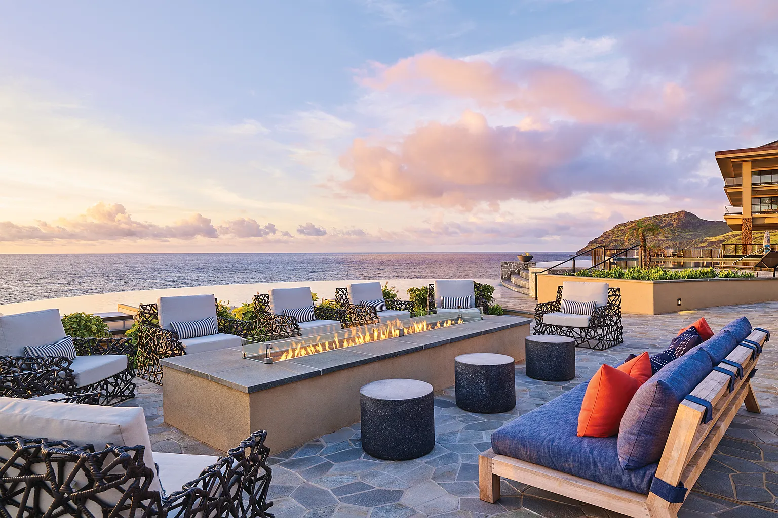 This inviting outdoor patio boasts a luxurious setting with comfortable seating arranged around a modern fire pit. The space offers stunning views of the ocean and a tranquil sunset, creating a desirable ambiance for relaxation and entertainment. Stone flooring and tasteful landscaping enhance the overall appeal of this upscale outdoor living area.