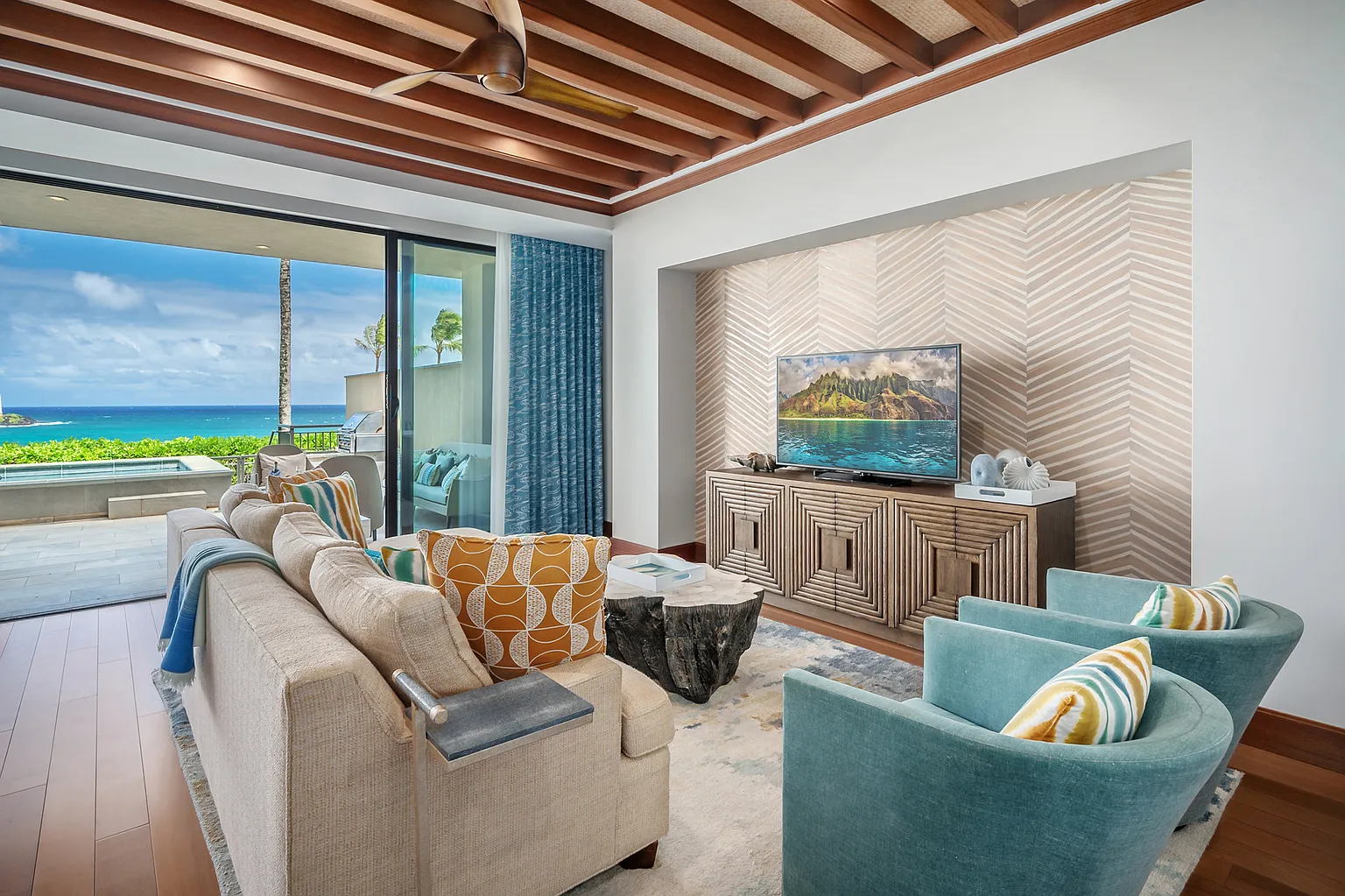 This is a beautifully designed living room with a stunning ocean view. The room features a comfortable sofa, two teal armchairs, and a unique coffee table, along with a well-placed TV above a modern entertainment center. The combination of natural light, luxurious furnishings, and stylish decor creates an inviting and sophisticated atmosphere.