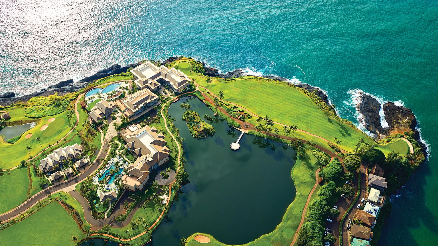 This aerial view showcases a luxurious coastal property with a well-manicured golf course, a large pond, and upscale residences. The image highlights the prime location along the oceanfront, featuring stunning turquoise waters and rocky cliffs. The development exudes exclusivity and high-end resort living.
