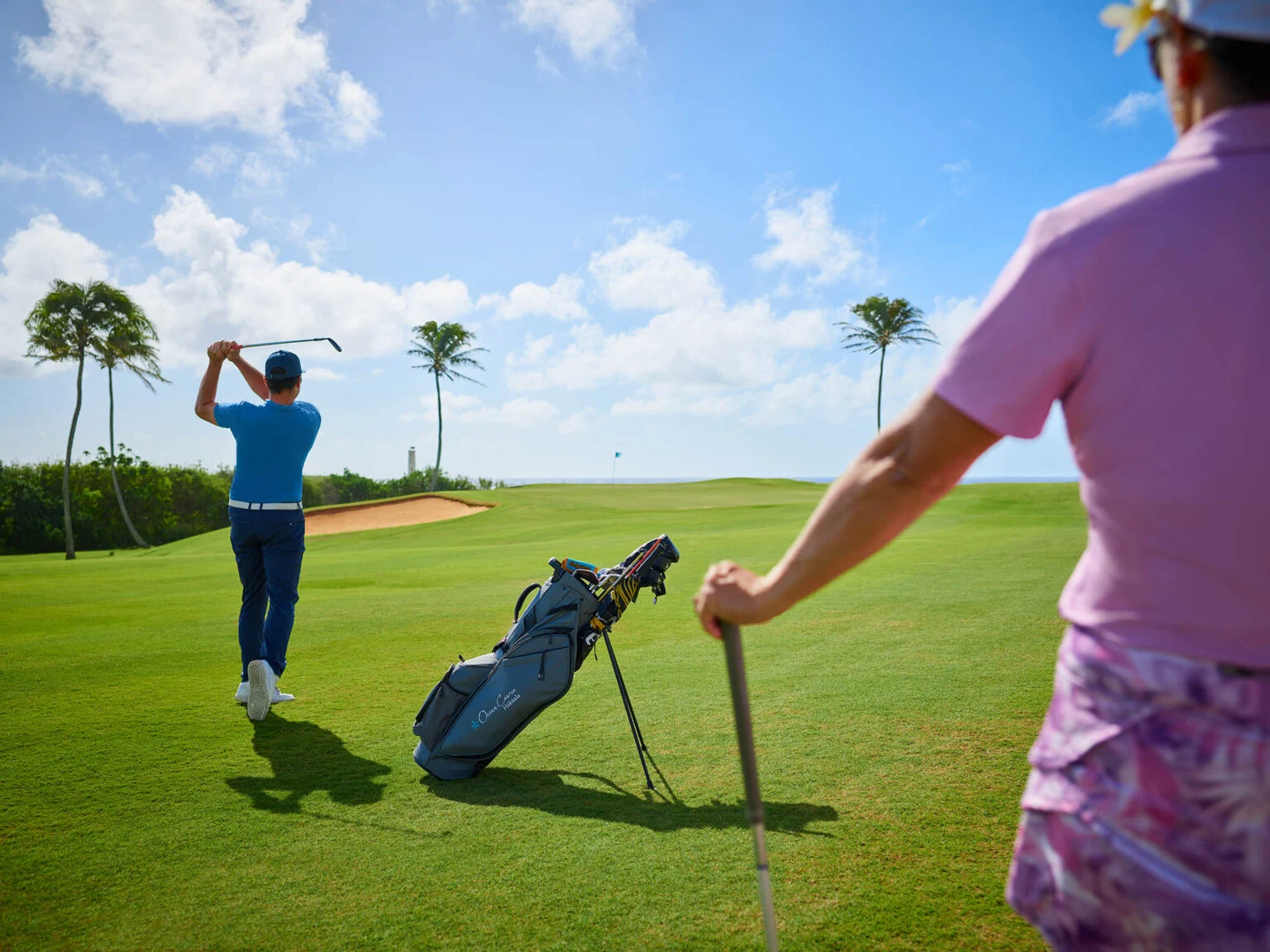 This image shows a view of a golf course, likely associated with a residential community or resort. The well-manicured green, sand traps, and palm trees suggest a high-end amenity. A golfer is seen in the distance, while another person with a golf club stands in the foreground, indicating the active lifestyle offered by the community.