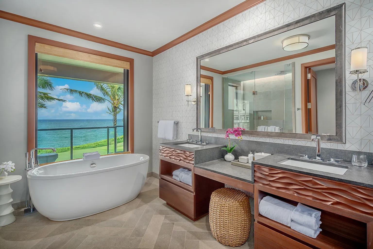 This is a luxurious primary bathroom featuring a freestanding soaking tub positioned next to a large window with ocean views. The design includes a double vanity with uniquely textured cabinetry, a large mirror reflecting the shower area, and stylish fixtures. The space exudes tranquility and high-end comfort.