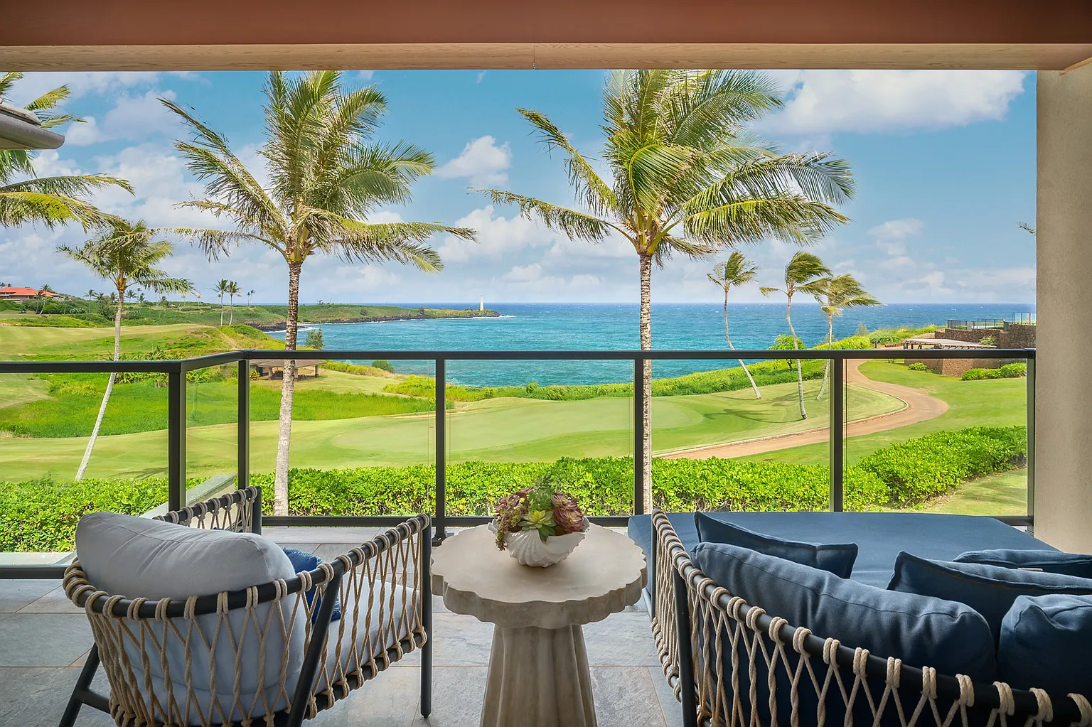 This image showcases a luxurious balcony or patio with comfortable seating. The space is framed by a glass railing, offering an unobstructed view of a lush green landscape with palm trees and the ocean. The overall impression is one of relaxation and high-end resort living.