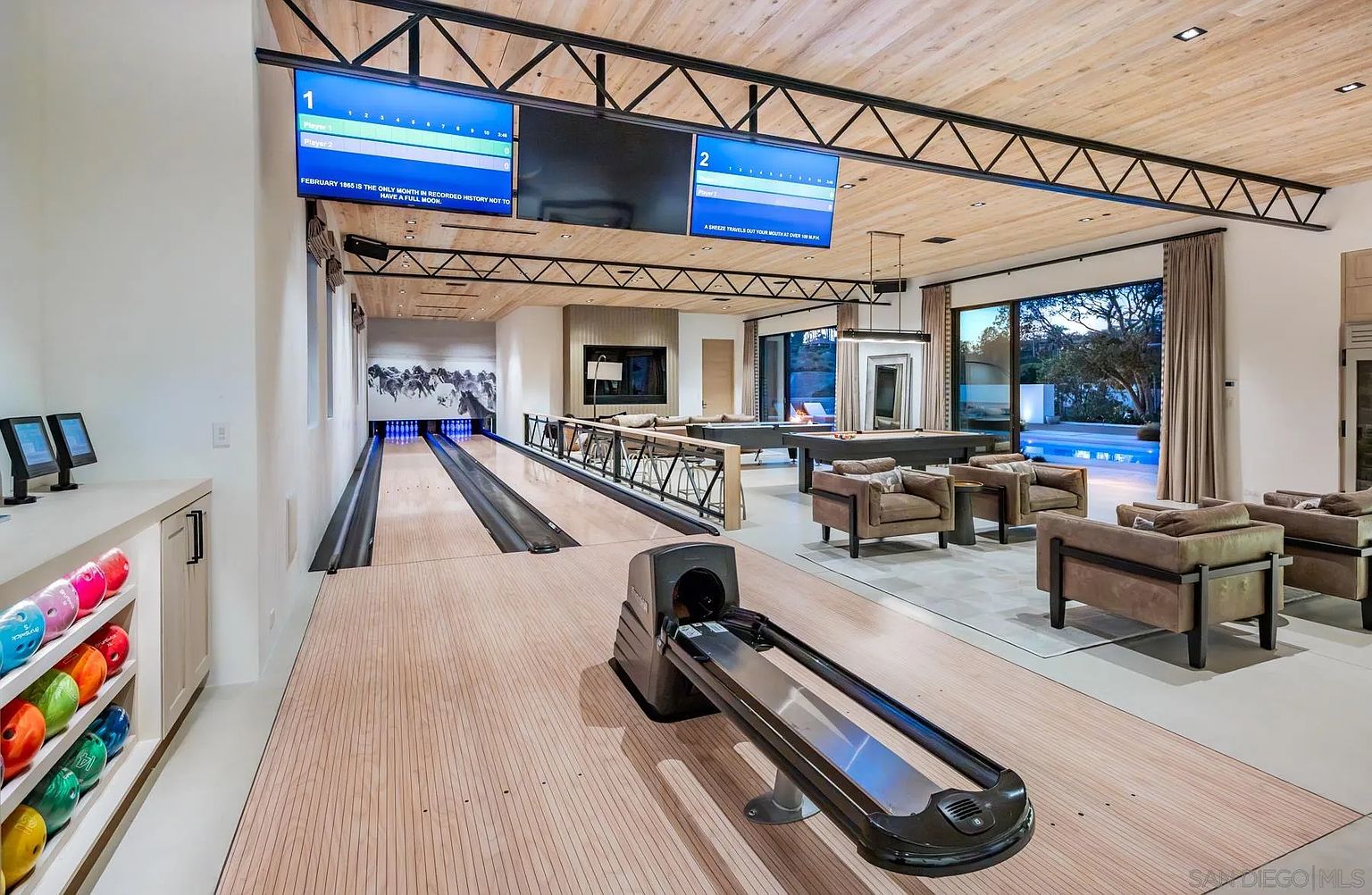 This expansive indoor entertainment space features a professional-grade bowling alley as its centerpiece, complete with ball returns and scoring monitors. The room transitions into a lounge area with a pool table and comfortable seating, all under a high, exposed-truss ceiling with light wood paneling. Large glass doors open to an outdoor pool area, creating a seamless indoor-outdoor flow perfect for hosting.