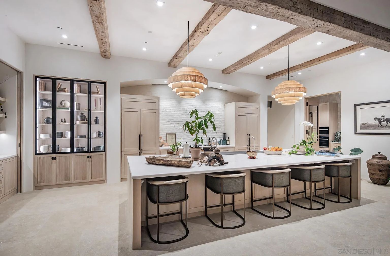 This sophisticated kitchen features a large central island with seating for five, topped with a light-toned surface and accented by two tiered, woven pendant lights. The space is defined by exposed wooden ceiling beams, light-colored cabinetry, and a white brick backsplash, creating a warm, transitional aesthetic. A tall, glass-fronted display cabinet with dark framing adds a striking visual contrast to the neutral palette, while the open-concept layout provides a clear view into the adjacent living area.