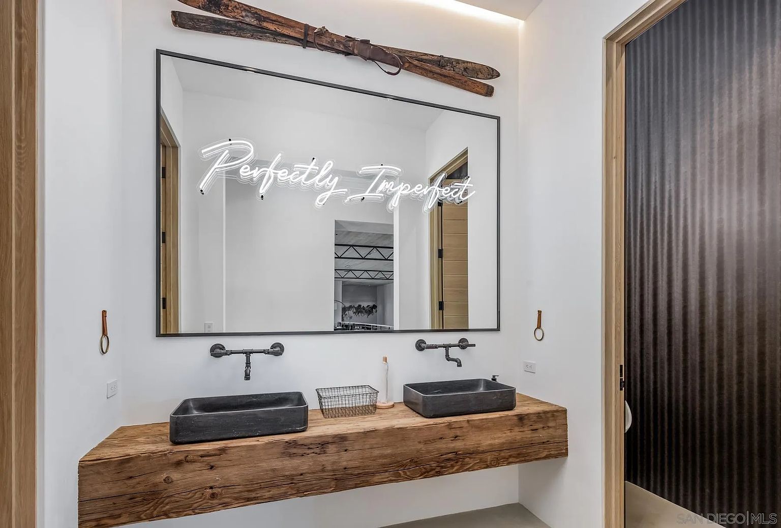 This modern bathroom features a rustic, thick-cut wood vanity topped with two dark, rectangular vessel sinks and wall-mounted industrial-style faucets. A large, rectangular mirror hangs above the vanity, adorned with a neon sign reading 'Perfectly Imperfect' and topped with a decorative pair of vintage wooden skis. The space is framed by clean white walls and a textured, dark-paneled door, creating a unique blend of mountain-inspired charm and contemporary design.
