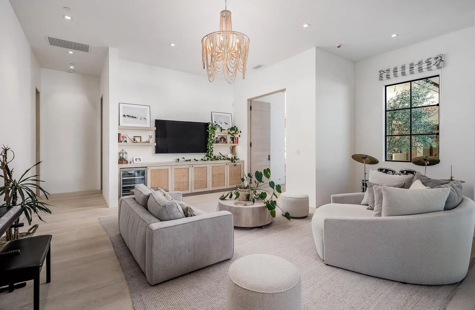 This bright and airy living room features a contemporary design with light wood flooring and a neutral color palette. The space is anchored by a large, curved sofa and a matching rectangular sofa, centered around a round coffee table and two textured ottomans. A media console with cane-front cabinetry sits beneath a wall-mounted television, complemented by open shelving and a beaded chandelier that adds a touch of organic texture to the room.