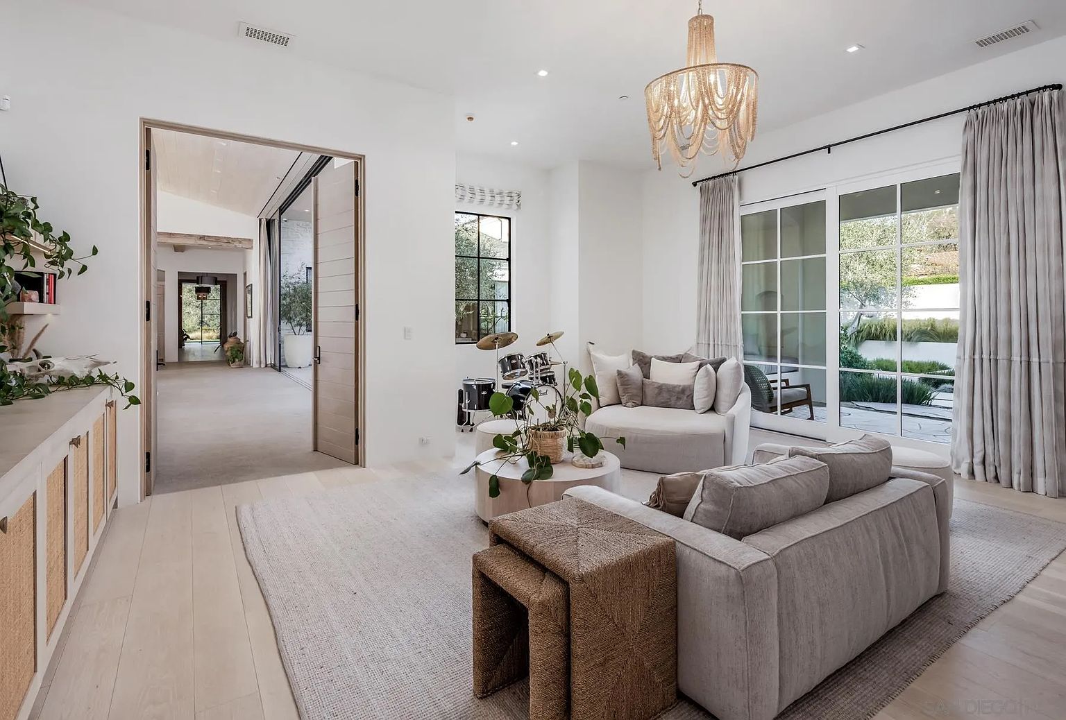 This bright and airy living room features a neutral color palette with light wood flooring and a large area rug. The space is furnished with a plush sofa, a circular coffee table, and a curved accent chair, while a drum set sits near the window. Large glass doors with floor-to-ceiling curtains open to an outdoor area, and a beaded chandelier hangs from the ceiling, adding a touch of texture to the room.