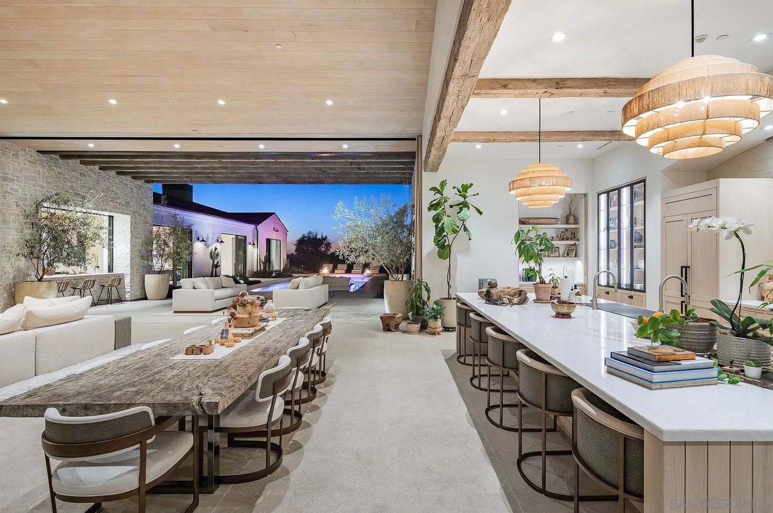 This expansive, open-concept living space seamlessly blends a modern kitchen with a dining area, featuring a large island with bar seating and a rustic, long wooden dining table. The room is defined by light wood ceilings, exposed timber beams, and a neutral color palette that creates an airy, sophisticated atmosphere. A massive opening leads directly to an outdoor patio and pool area, emphasizing a fluid indoor-outdoor lifestyle perfect for entertaining.