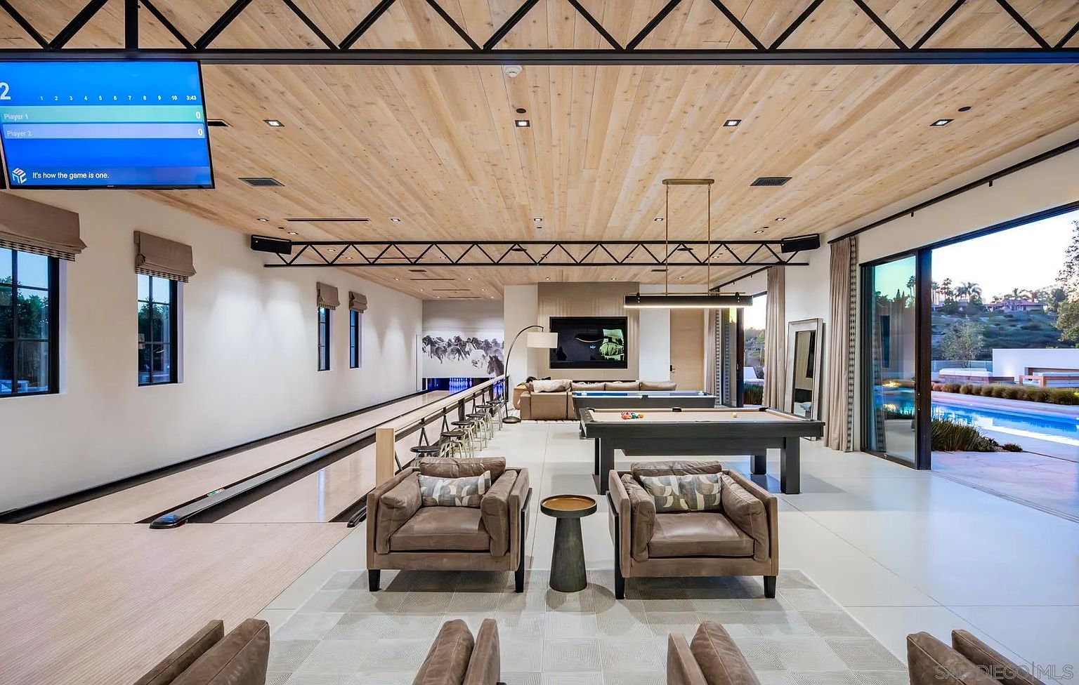 This expansive, modern entertainment room features a private bowling alley on the left and a billiards area on the right, creating a luxurious recreational space. The room is defined by a light wood-paneled ceiling with exposed black metal trusses, large glass sliding doors that open to an outdoor pool area, and comfortable leather seating in the foreground. The aesthetic is sleek and contemporary, blending high-end leisure amenities with an open-concept design.