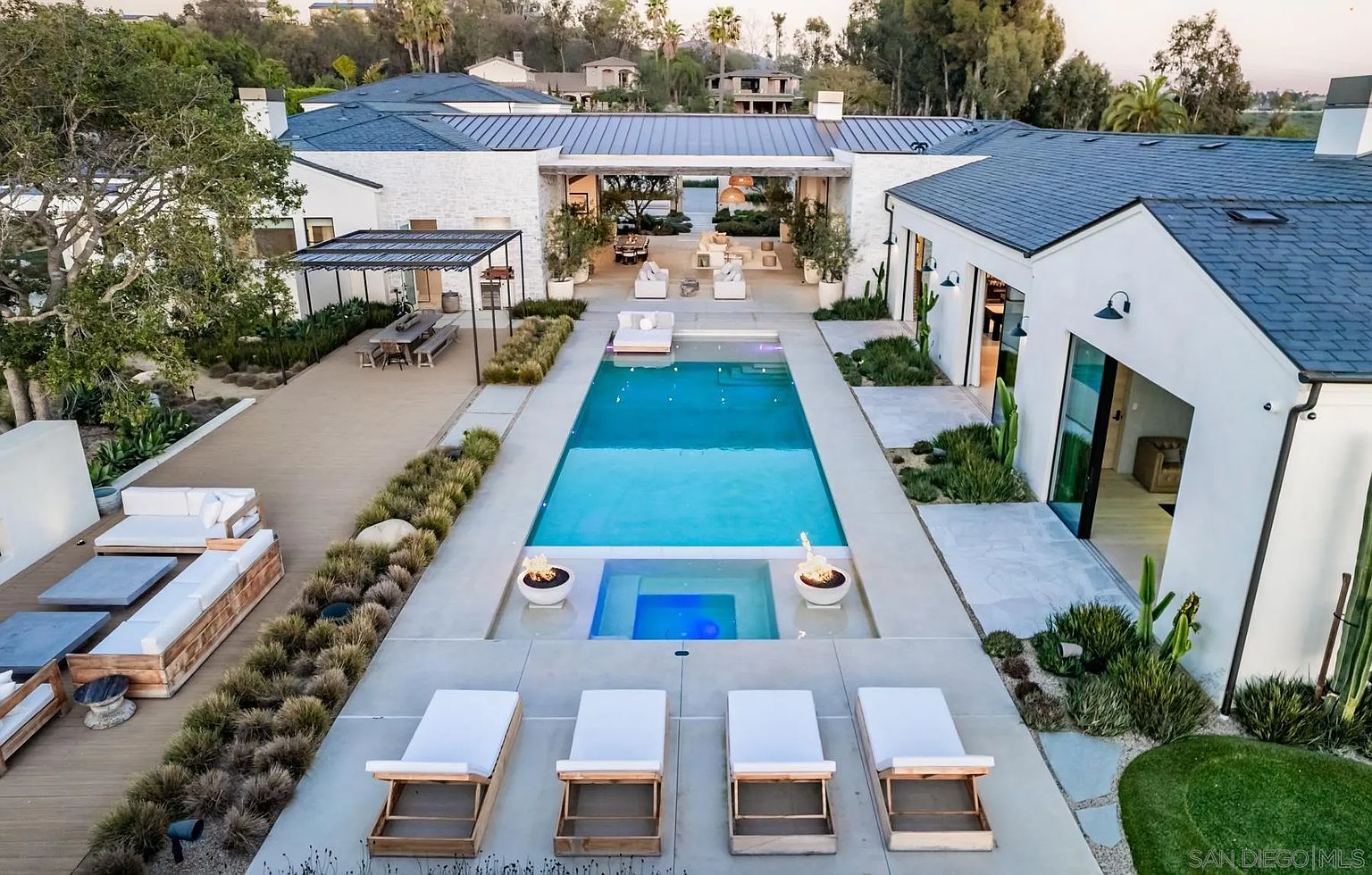 This high-angle aerial view captures a luxurious modern residence centered around a sprawling rectangular swimming pool and integrated spa. The property features a symmetrical layout with white exterior walls, dark metal roofing, and a covered outdoor living area that seamlessly connects the two wings of the home. The meticulously landscaped grounds include a wooden deck with a dining area, multiple lounge spaces with white furniture, and fire features flanking the spa, creating a sophisticated and inviting resort-style atmosphere.