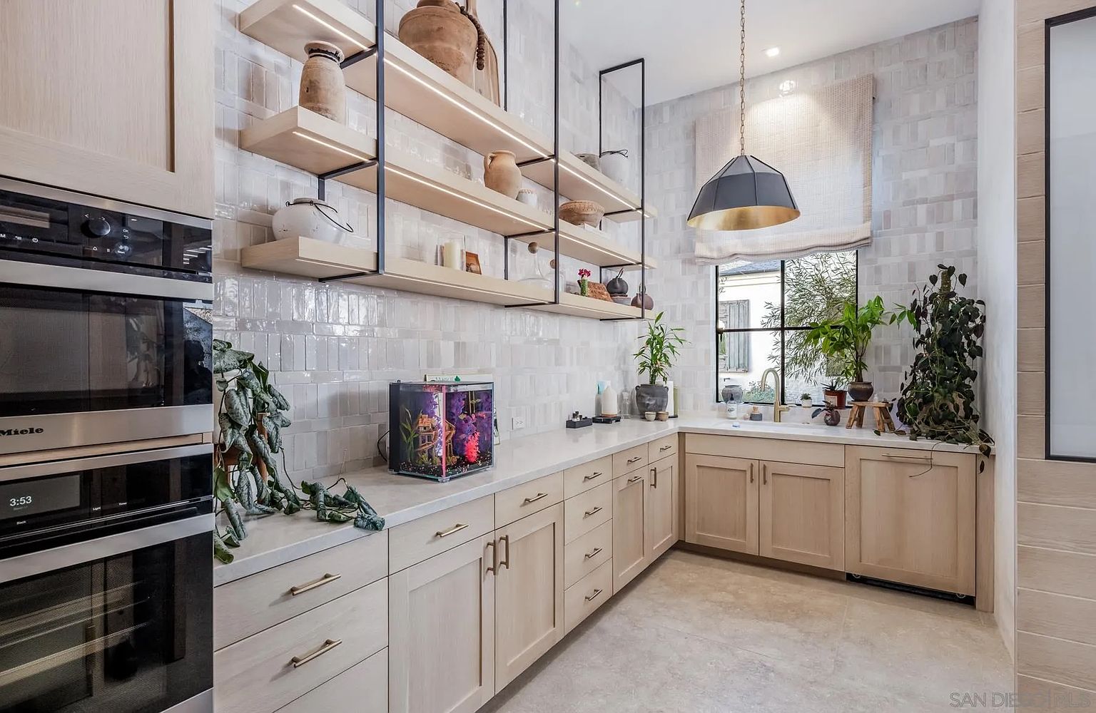 This bright and modern kitchen features light wood cabinetry paired with light-toned stone countertops and a textured tile backsplash. Floating shelves with integrated lighting are mounted on the wall, providing an open display area, while a large pendant light hangs above the sink area. The space is accented with various potted plants and a small aquarium, creating a warm and inviting atmosphere.