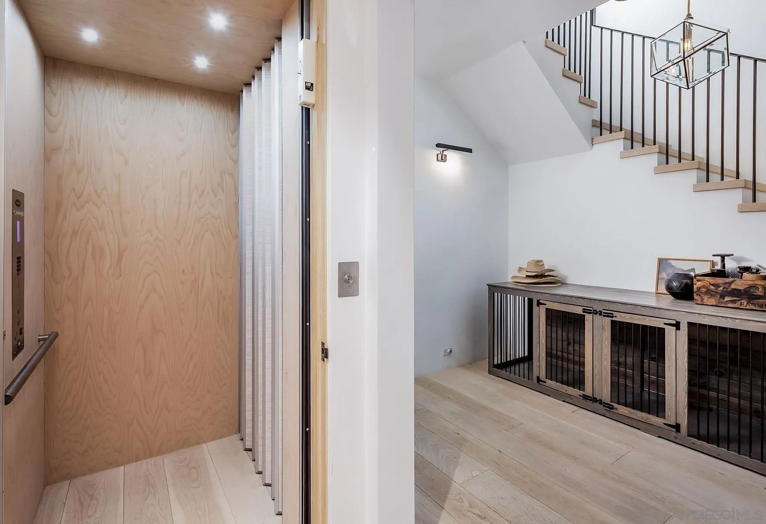 This interior view captures a modern home elevator with light wood paneling and a folding door, positioned next to a hallway leading to a staircase. To the right, a custom-built wooden console with integrated pet crates sits beneath a staircase featuring open risers and a sleek metal railing. The space is illuminated by recessed lighting and a geometric pendant fixture, creating a clean, contemporary aesthetic.