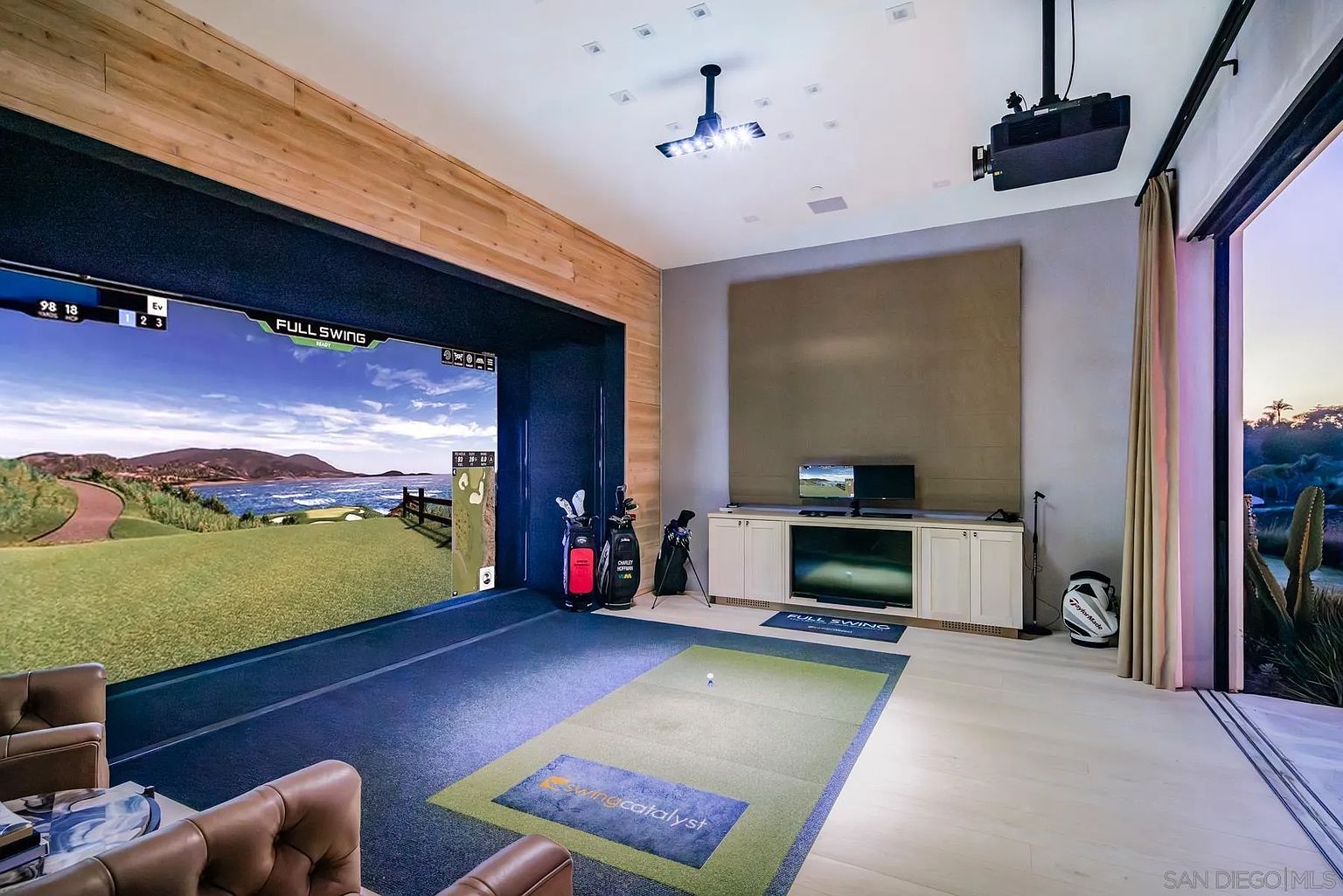 This image showcases a high-end indoor golf simulator room, featuring a large projection screen displaying a virtual course and a dedicated putting mat with integrated technology. The space is designed for entertainment, with a built-in media console, golf bag storage, and a comfortable leather seating area in the foreground. Large sliding glass doors open to an outdoor view, blending indoor recreation with the surrounding landscape.